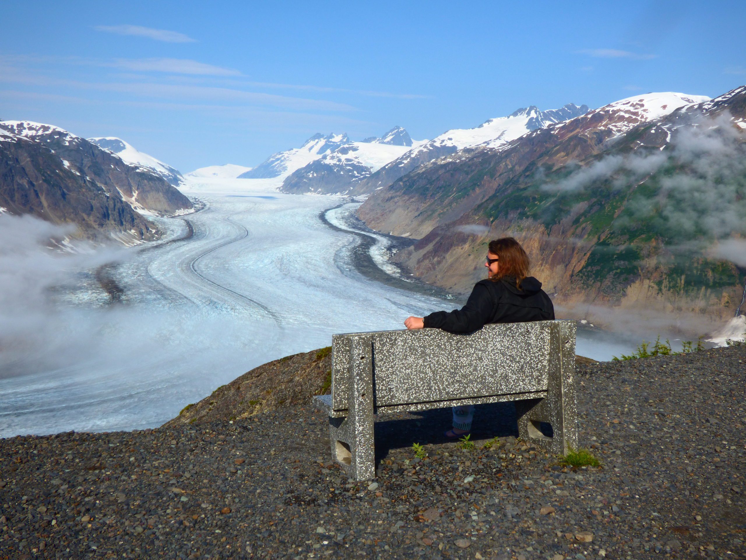 Woman sits on concrete bench overlooking Alaskan glacier