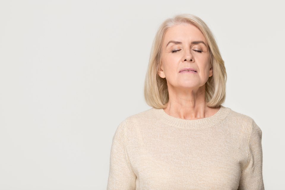 Calm tranquil mid aged old woman meditating breathing fresh air, serene mindful mature senior lady taking deep breath feeling no stress free relief harmony isolated on white grey studio background