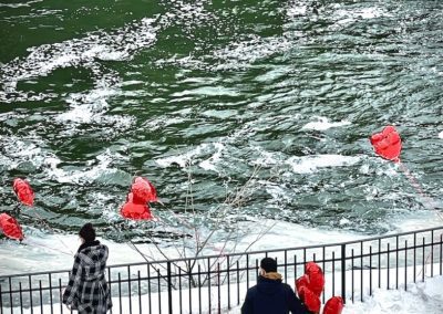 A man looks at a woman, both holding red heart-shaped balloons next to a body of water