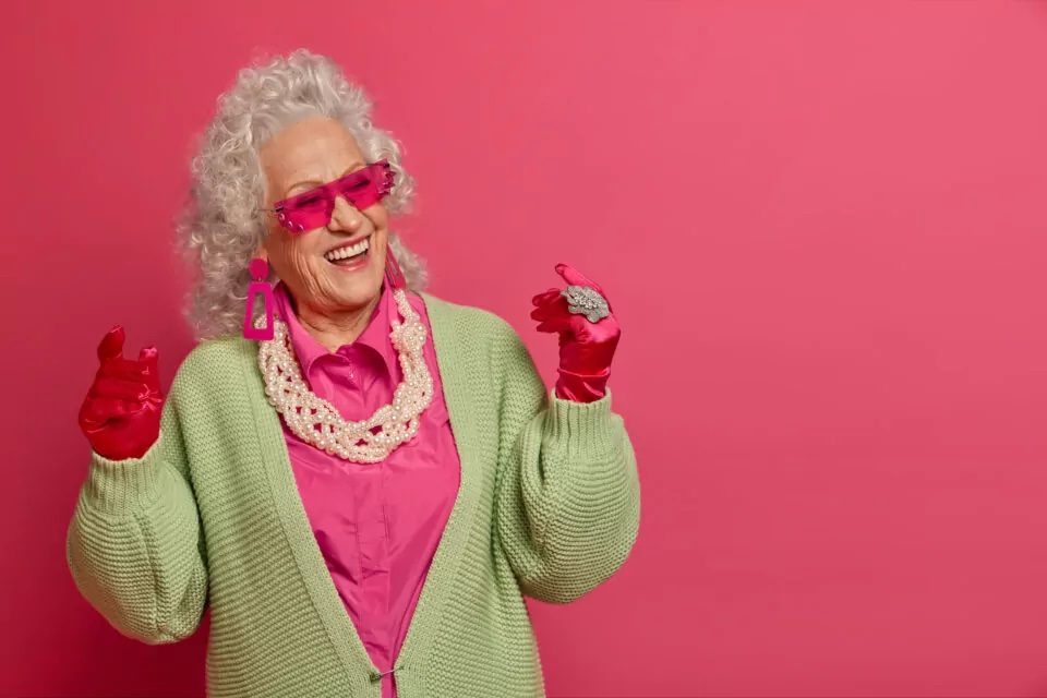 Happy senior woman wears pink gloves, glasses, and lots of jewelry against a pink background