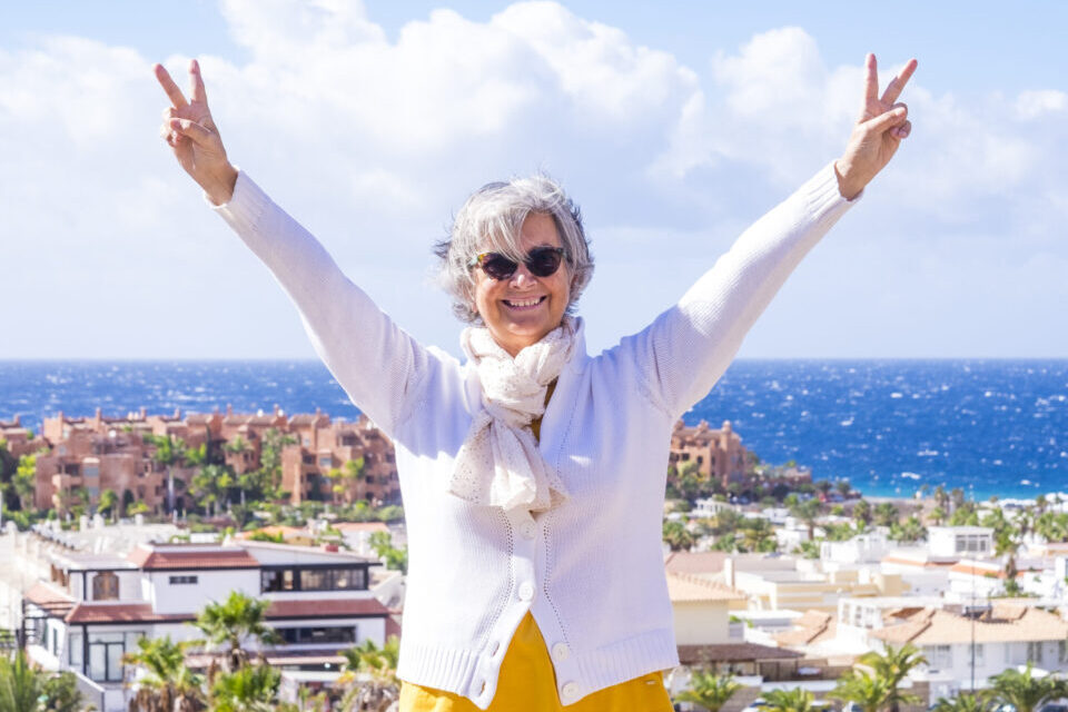 Happy senior woman enjoying sea holiday in winter. Positive, optimistic active retiree smiling with arms up