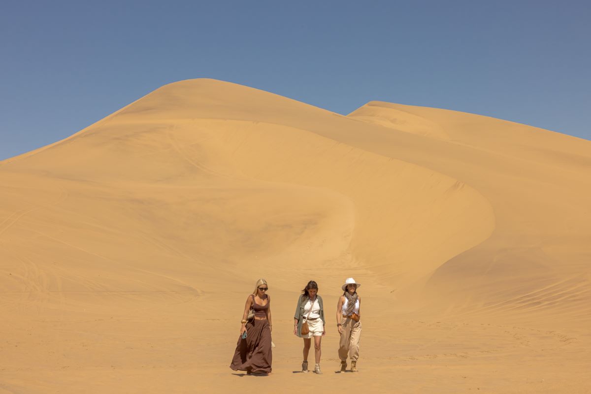 three ladies standing in front of giant sand dunes against a blue sky backdrop
