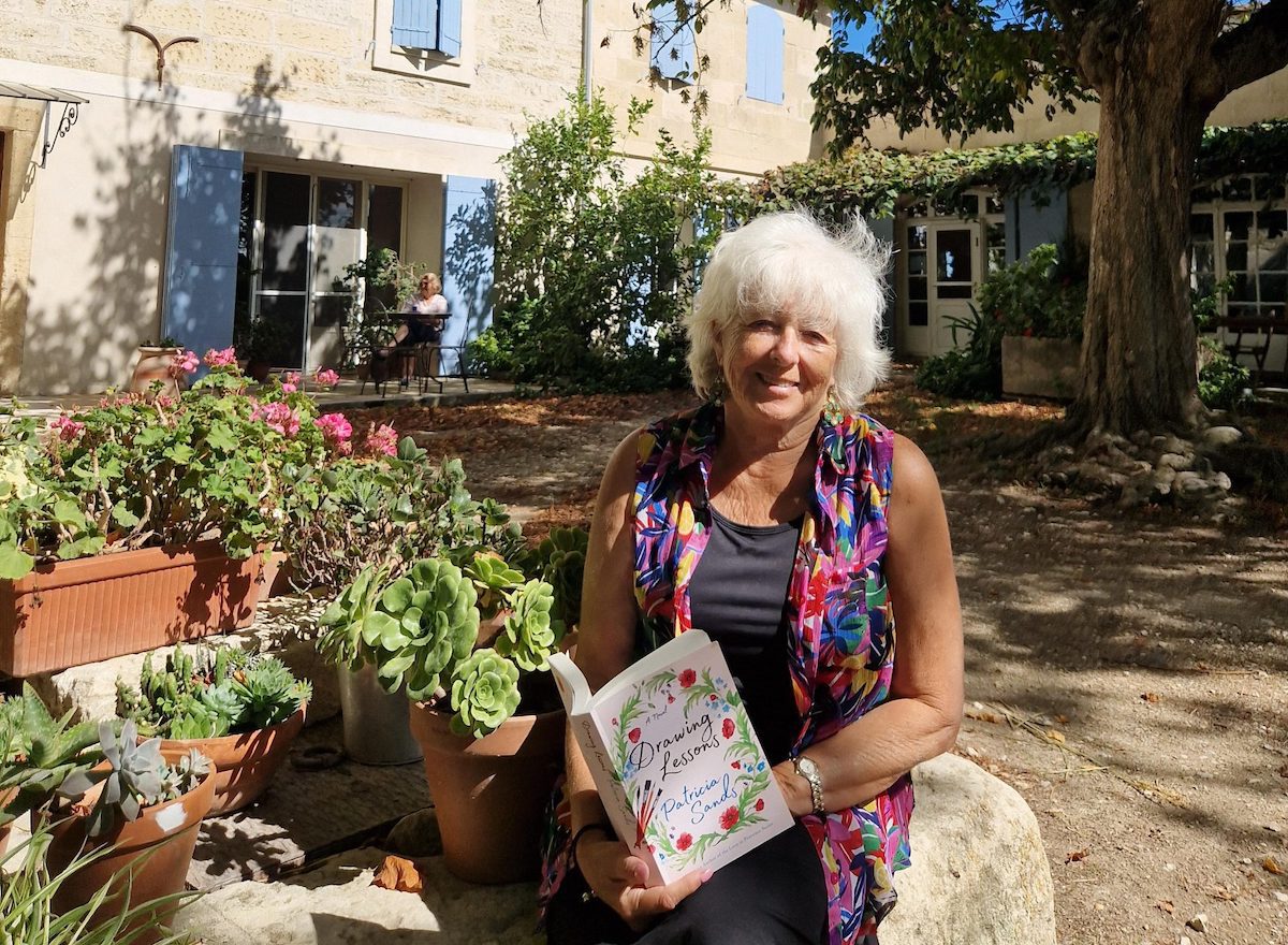 Author Patricia Sands, sitting in front of a provencal house, holding up one of her novels.