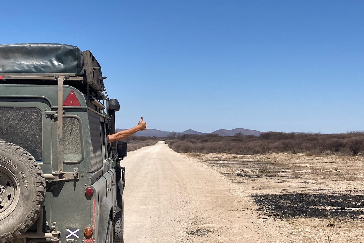 A vehicle drives down a dirt road while the passenger gives thumbs up out the window during a road trip across southern Africa.