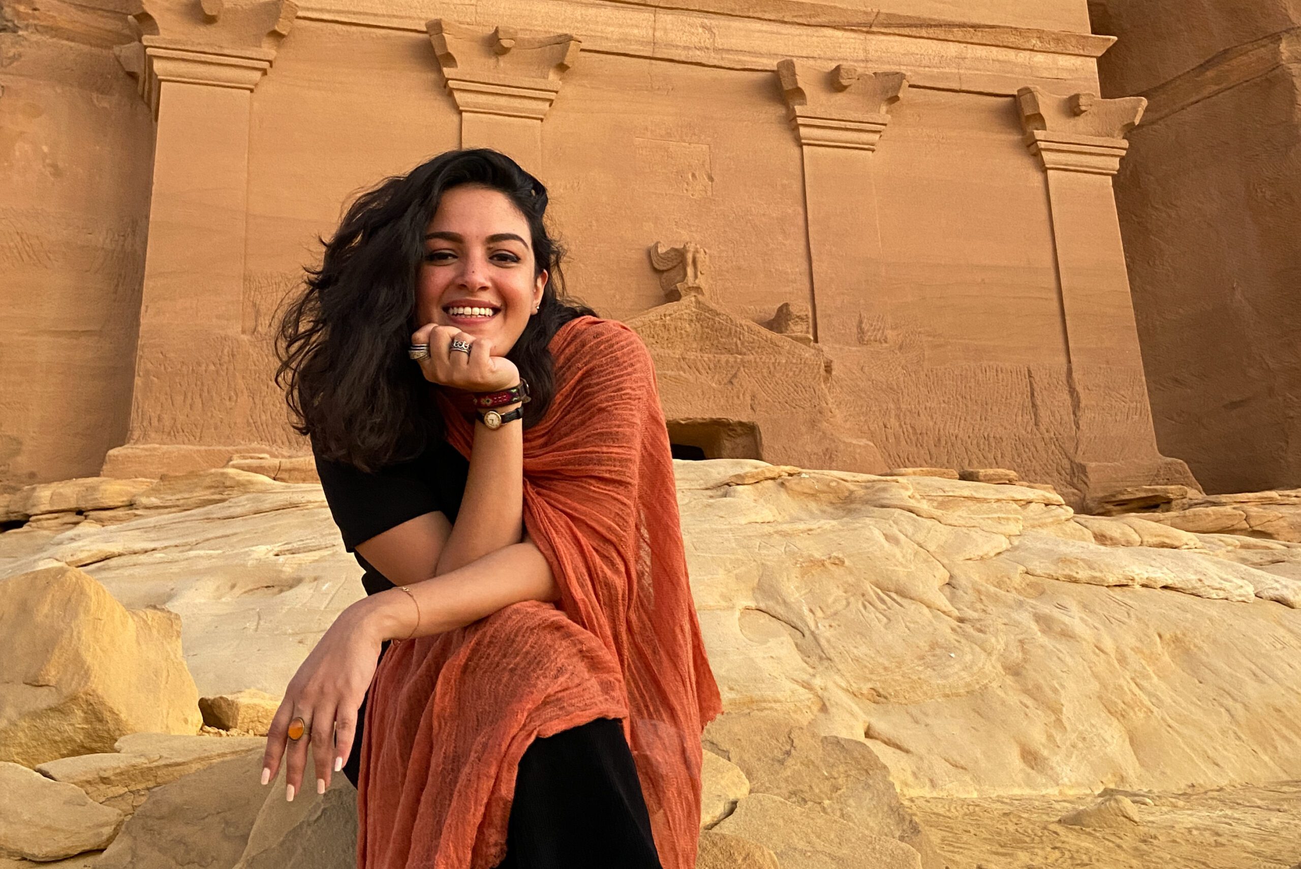 Woman sitting down in front of a tomb smiling and wearing an orange shawl