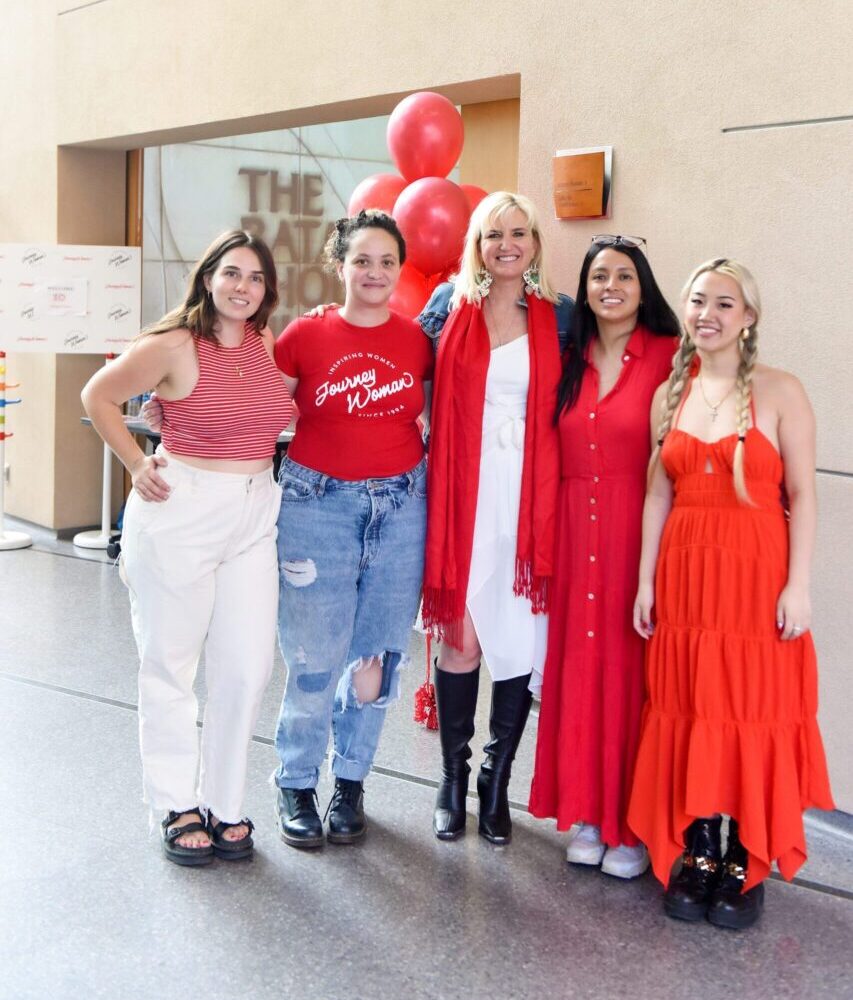 five women wearing red smiling in a semi circle at event with balloons in the background