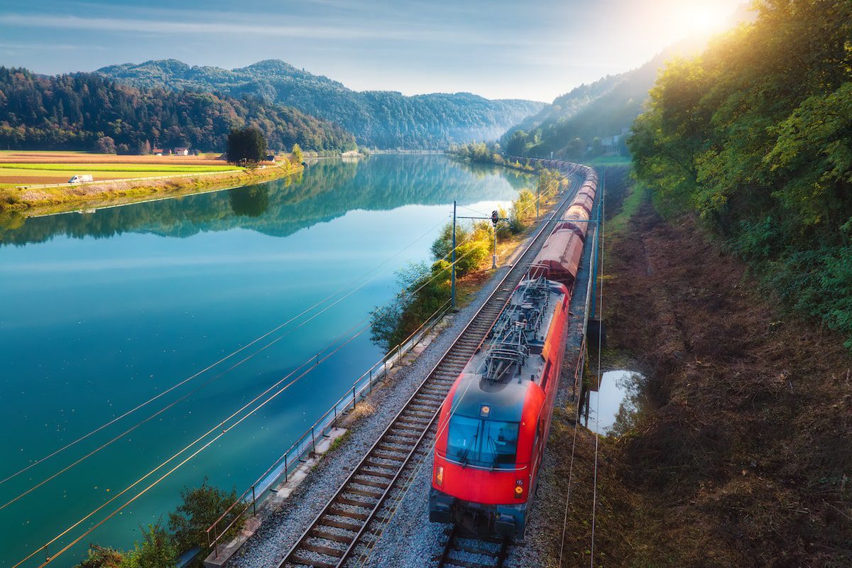 Aerial view of red modern high speed train moving near river in alpine mountains at sunrise in spring