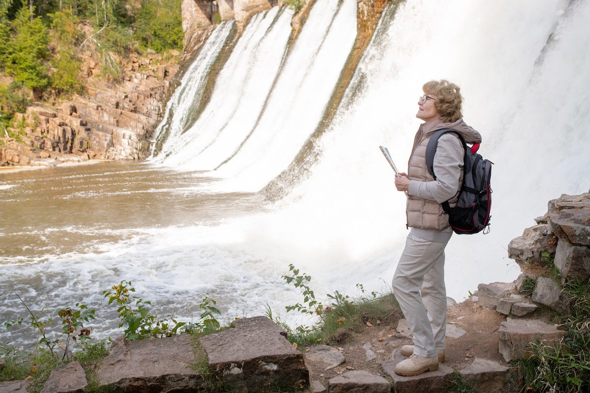 Mature female hiker with backpack and map guide standing by waterfalls in natural environment and seeking right way