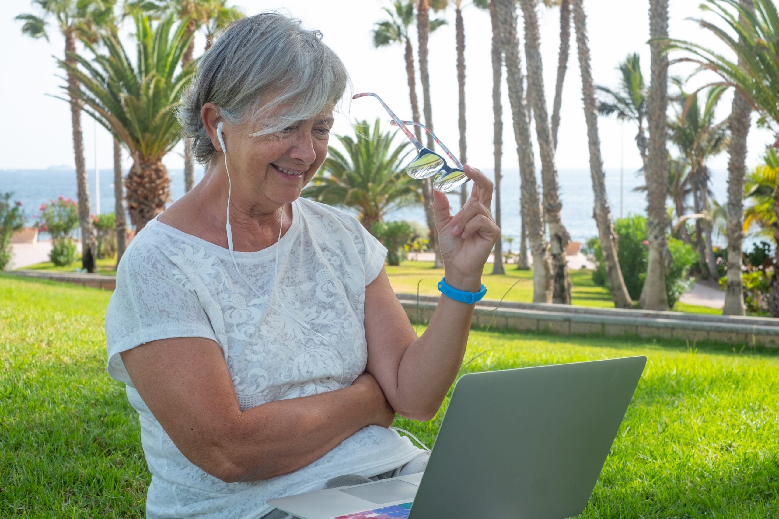 Smiling senior woman with gray hair sitting in the grass with the laptop ready for online purchases By lucigerma skyscanner