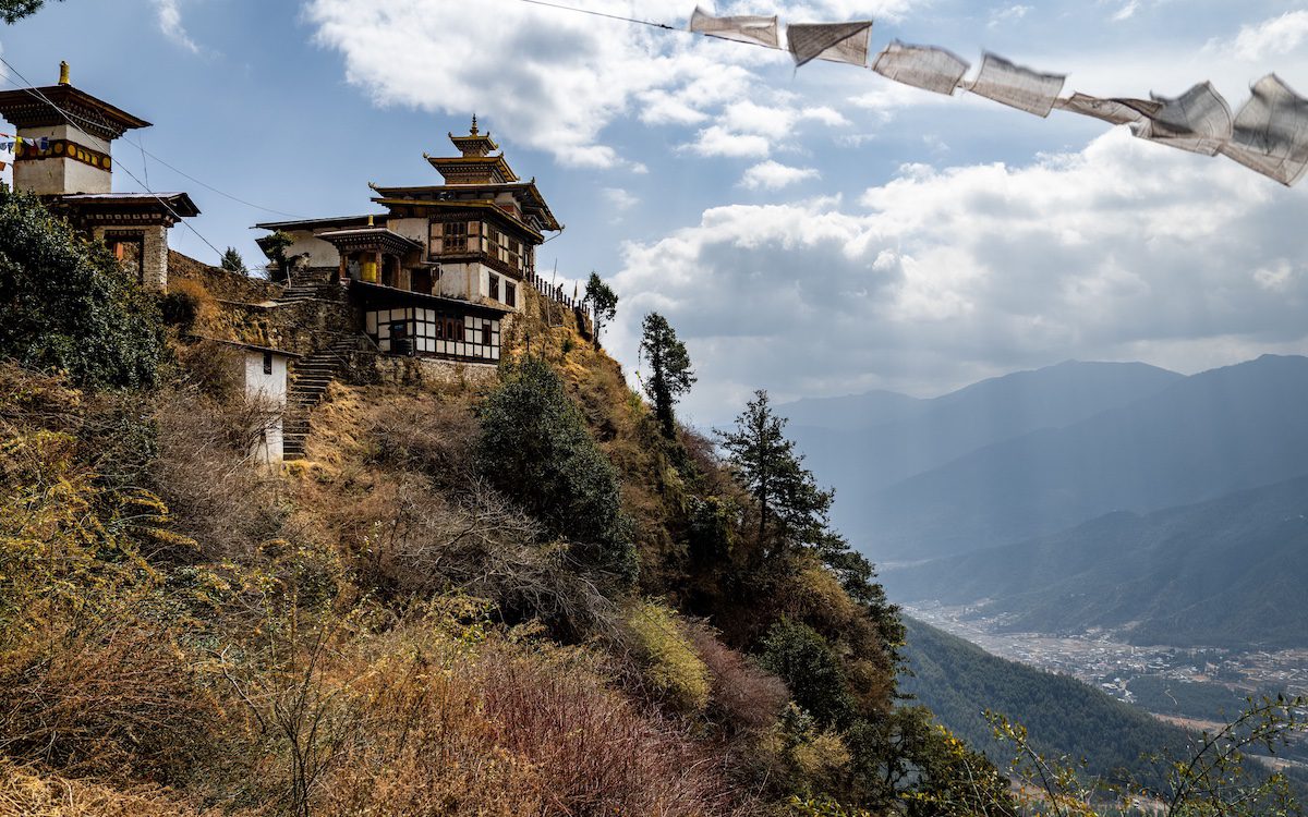 Traditional Bhutanese monastery perched on a hill with prayer flags and scenic valley view under a cloudy sky.