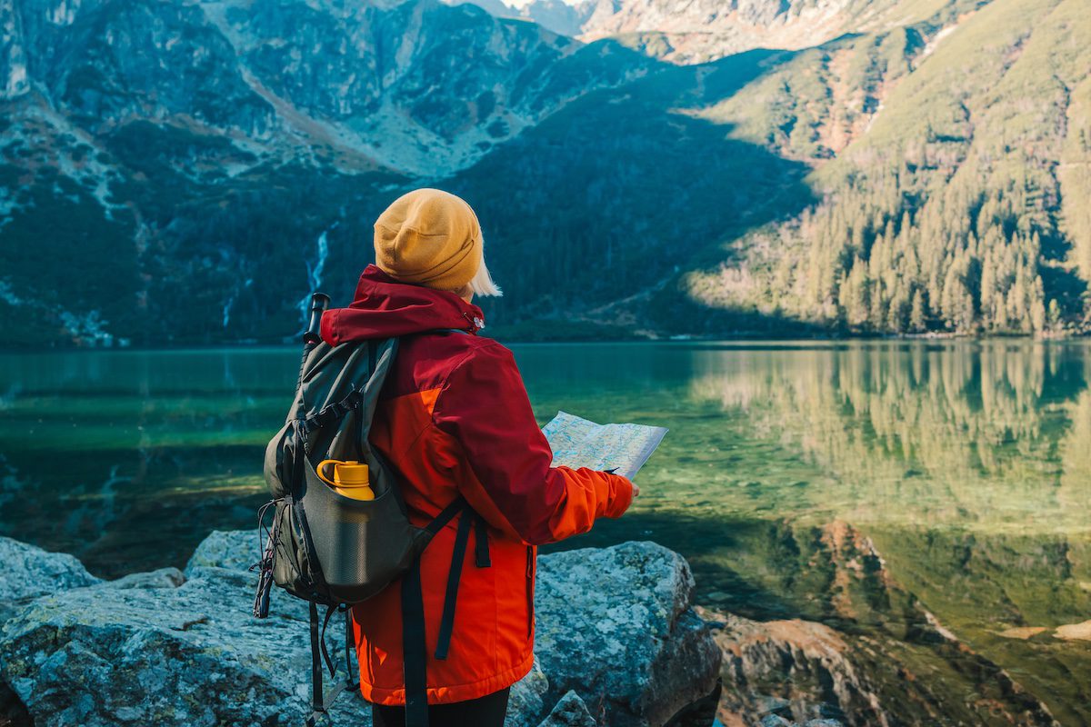 Shot of a senior 60s lost woman with smart phone holding a map, learning how to decide where to travel, while taking in the view from the top of a mountain. Female tourist sits on the rock in mountain read the map. Poland, Tatry