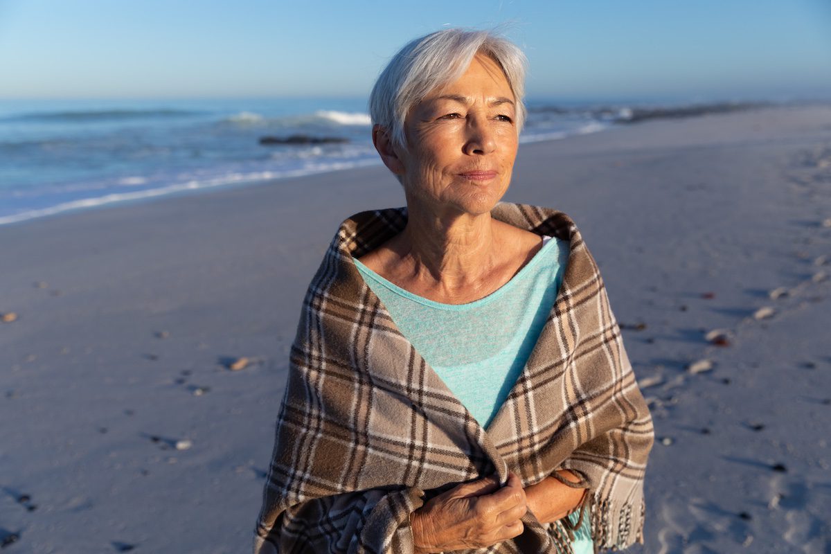 Senior woman enjoying time at the beach, standing with a blanket around her shoulders